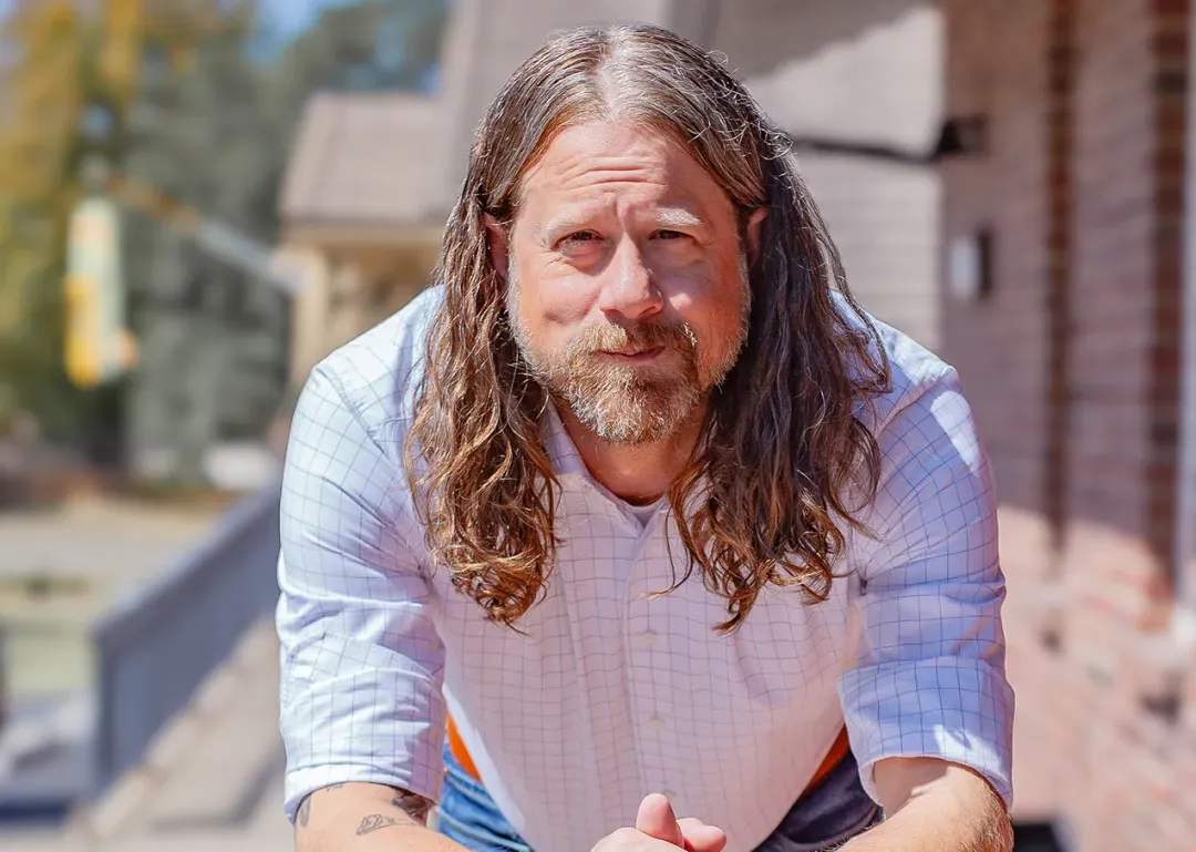 Man with long hair and beard leaning forward outdoors near a brick building.