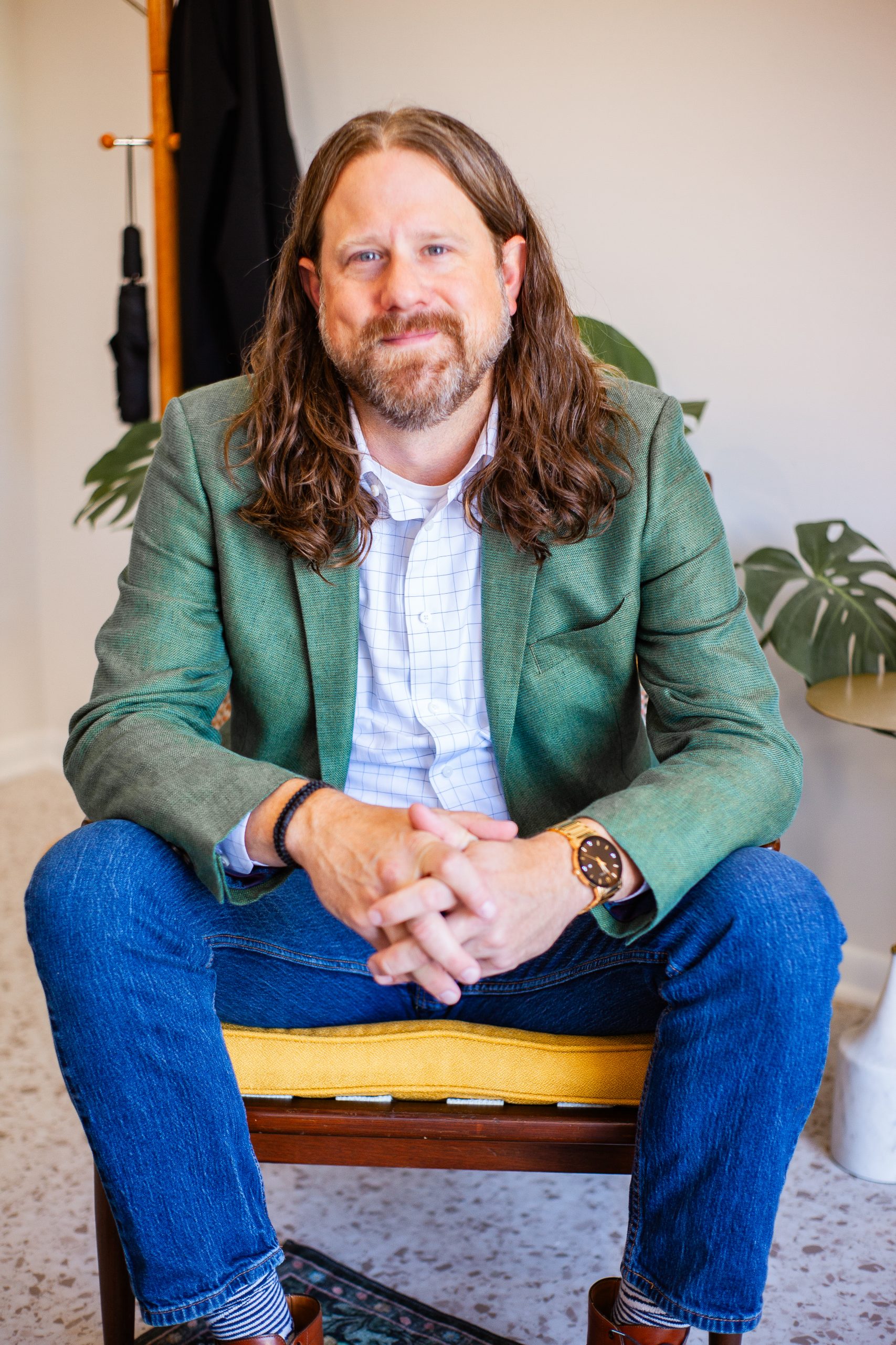 Man with long hair and a beard sitting cross-legged wearing a green blazer and blue jeans.