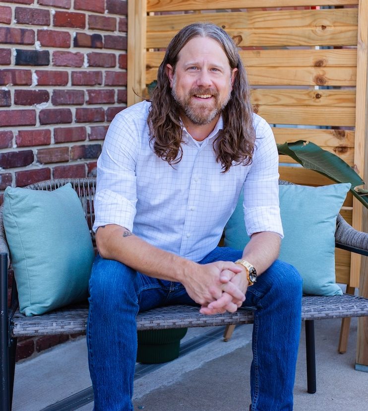 Man with long hair and beard wearing a white checkered shirt leaning forward outdoors.