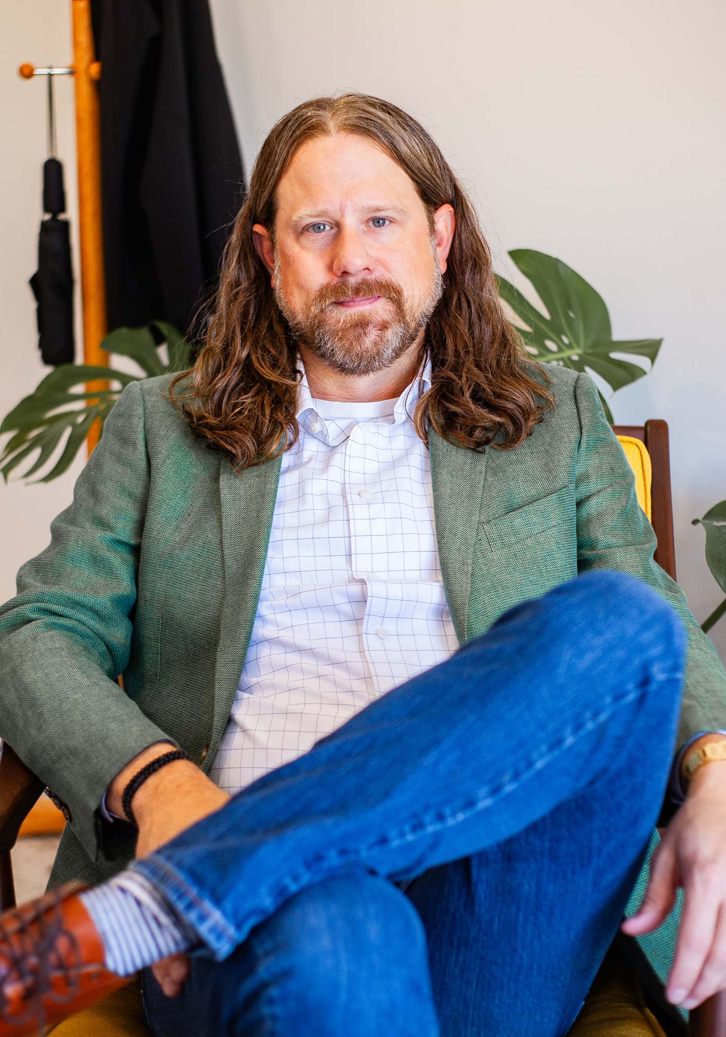 Man with long hair and beard sitting cross-legged on a yellow chair wearing a green blazer, white checkered shirt, and blue jeans.